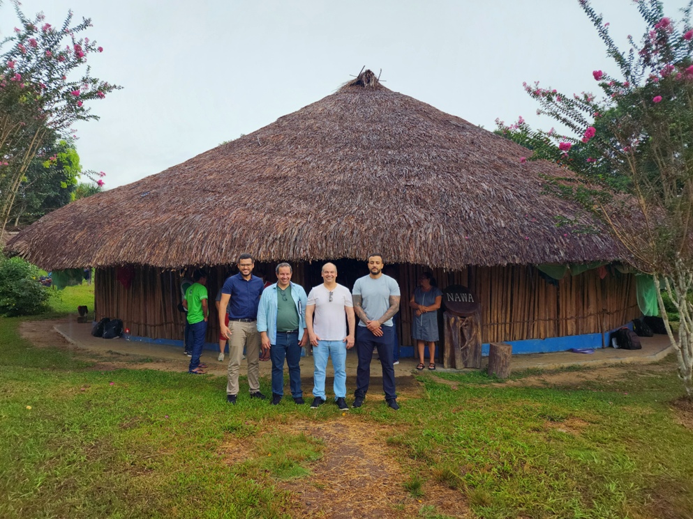 A imagem mostra um malocão na comunidade Waimiri Atroari, à frente, posando para a foto estão o coordenador do Cejusc, Eduardo Álvares Carvalho, o juiz titular da comarca de Rorainópolis, Phillip Barbeiux, o juiz auxiliar da terceira vice-presidência do Tribunal de Justiça de Minas Gerais (TJMG), Marcus Vinicius Mendes, e o juiz da comarca de Aguas Formosas de Minas Gerais, Matheus Moura .