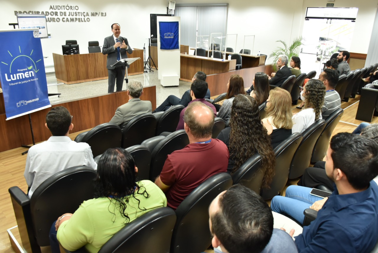 Imagem colorida mostra  o presidente TJRR, desembargador Cristóvão Suter, discursando em um auditório para um grupo de homens e mulheres, a foto mostra também, dois banners azuis, um com a logo do TJRR e outra com a logo do projeto lumen, ambas estão posicionadas nas laterais à frente da plateia.