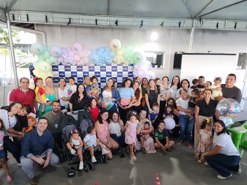 Imagem 1- Imagem colorida em formato retangular, mostra  pais, mães,  mães gestantes e  crianças entre  0 a 3 anos de idade, sorridentes. posando para a foto em frente a um painel decorado com balões coloridos