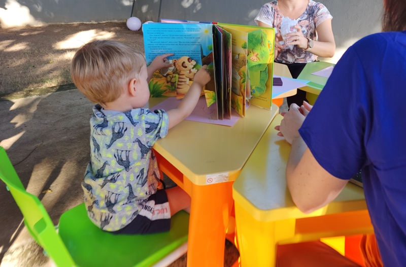 Imagem colorida mostra um menino com cabelos dourados sentado de costas para a foto,  em uma cadeira verde, folheando um livro infantil  sobre uma mesa amarela.