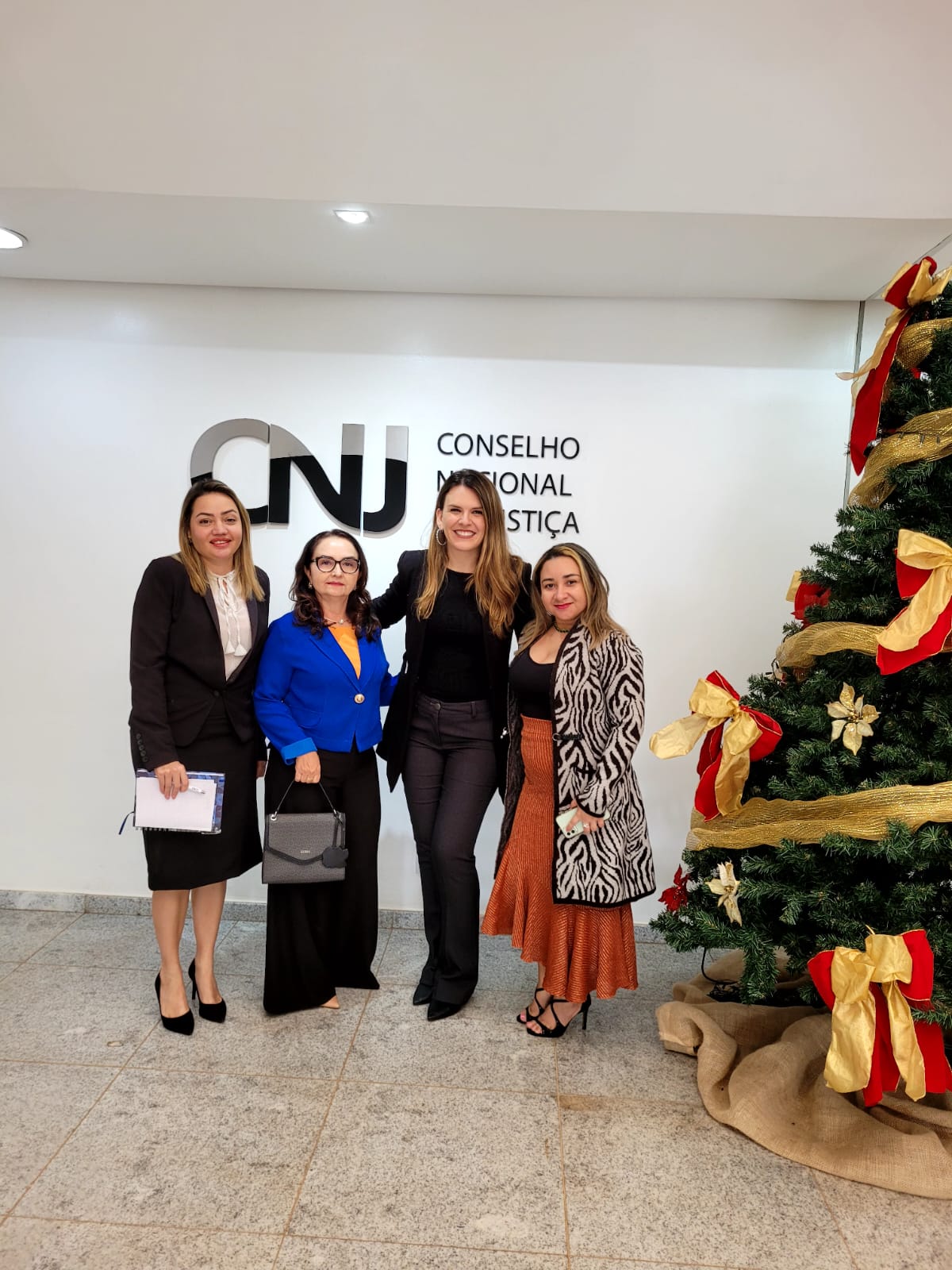  Imagem  colorida  em formato retangular, mostra as servidoras do cerimonial do Tribunal de Justiça de Roraima (TJRR), Haline Barreto, Domícia Marques e Solange Alencar, posando para a foto com representante das Secretarias de Cerimonial do Conselho Nacional de Justiça.