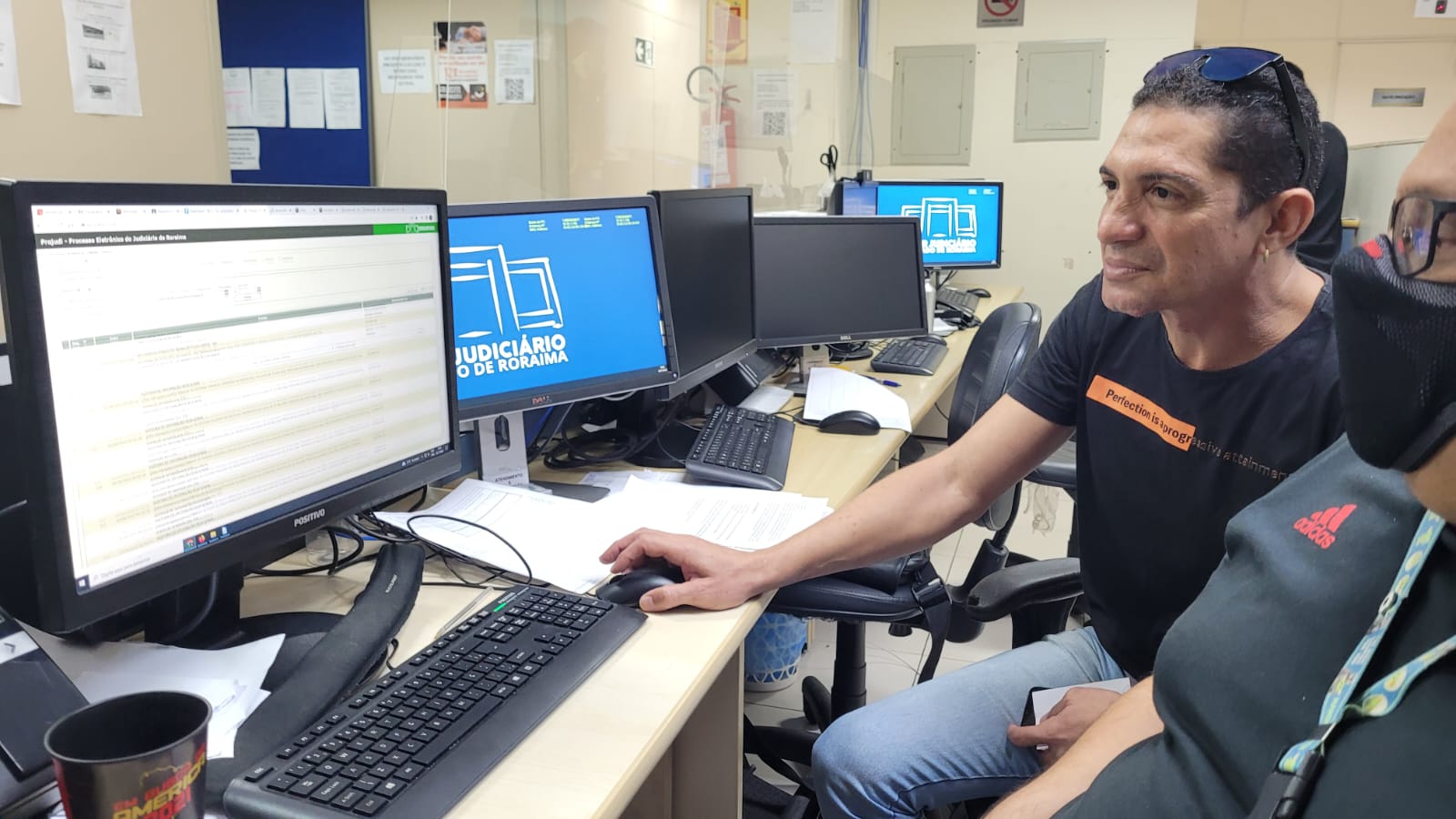 Imagem colorida  mostra o perfil de dois Servidores do Tribunal de Justiça de Roraima (TJRR), sentados em frente a uma mesa com cinco telas de computadores estando três telas ligadas, um dos servidores está segurando o mouse e fazendo a ação de clicar, em quando a outra observa a ação.