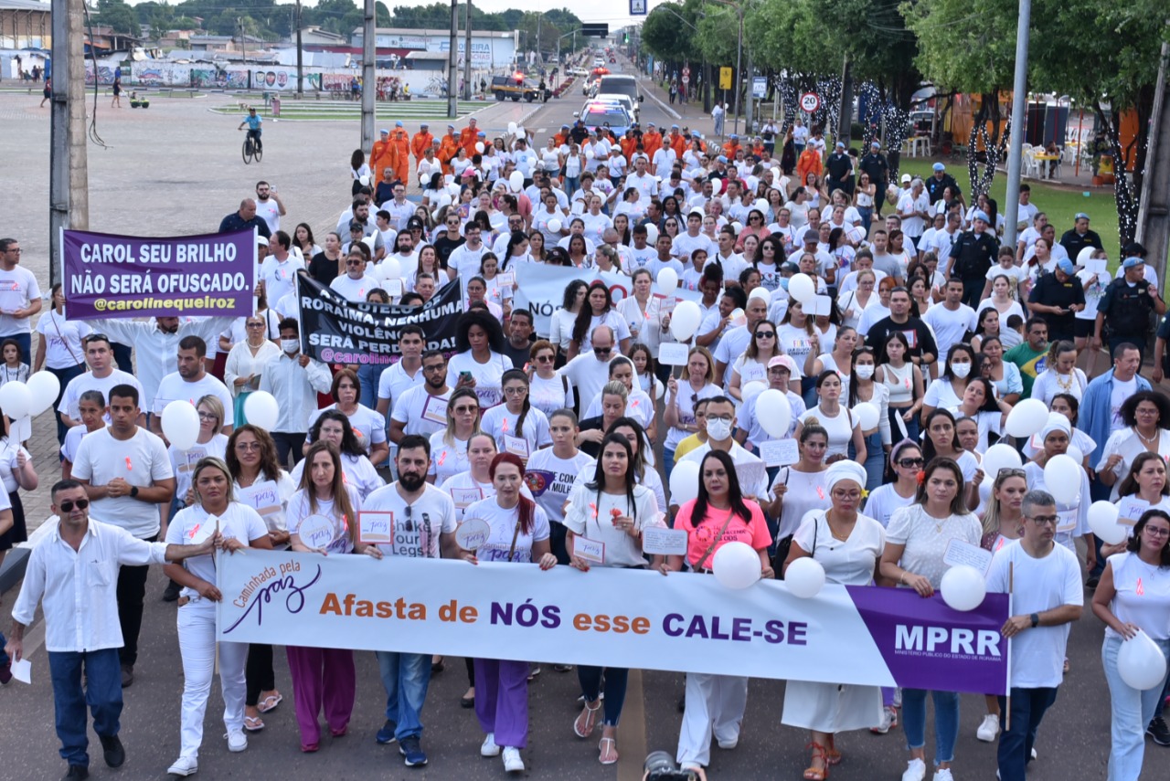 Imagem colorida em formato retangular, mostra homens e mulheres vestidos de branco e segurando cartazes  durante caminhada pela paz, o cartaz principal apresenta o texto “caminhada pela paz,  Afasta- se de nós esse cale-se, MPRR”   