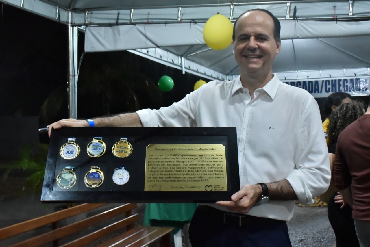 Imagem colorida em formato retangular, mostra o presidente do TJRR, desembargador Cristóvão Suter segurando a placa de homenagem dos servidores do Centro Médico e de Qualidade de vida.