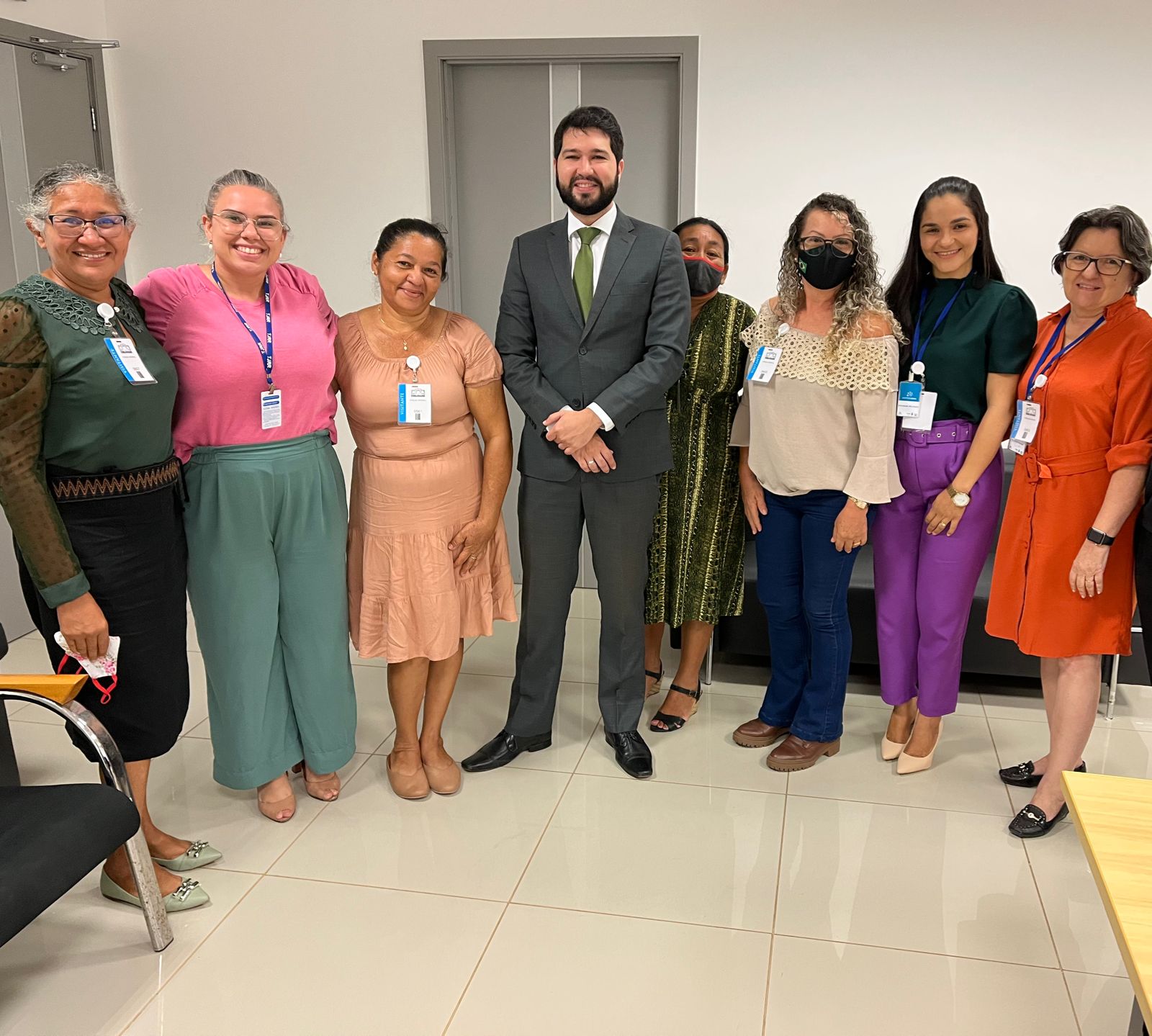  Imagem colorida em formato retangular mostra um homem de terno cinza e gravata verde ao lado de sete mulheres em pé posando para a foto.