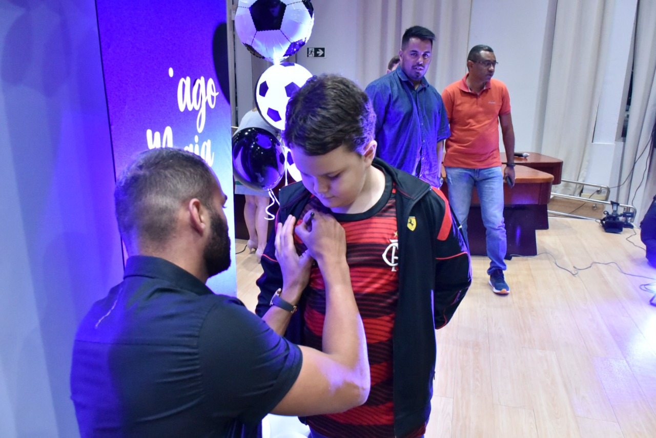 Imagem colorida em formato retangular mostra o jogador do Flamengo, Thiago Maia assinando a camisa de time de uma criança.