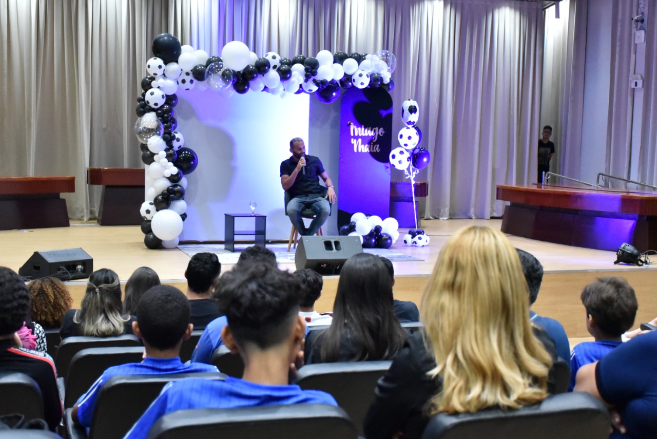 Imagem  colorida em formato retangular mostra o auditório do Fórum Cível Advogado  Sobral Pinto com um público de homens, mulheres, jovens, adolescentes e crianças durante a palestra “Ouro Norte”, ministrado pelo jogador do Flamengo, Thiago Maia, o mesmo estar a frente da plateia sentado , atrás há um painel decorado com balões nas cores preto e branco e eles  alguns balões  simulam bolas de futebol.