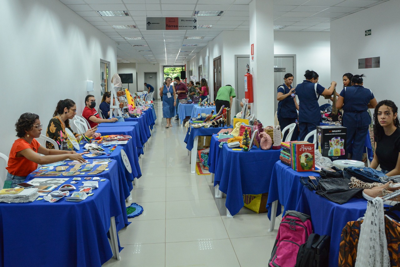 Imagem colorida em formato retangular, mostra uma feira de exposição de artesanato, com mesas cobertas com um pano azul, posicionadas em duas fileiras, sobre as mesas estão os trabalhos. Na foto há consumidores circulando e artesãos sentados por trás das mesas expondo seus respectivos trabalhos.