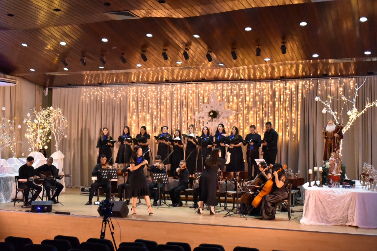 Imagem colorida em formato retangular mostra o coral formado pelos servidores do Poder Judiciário durante apresentação, o palco está enfeitado com luzes de natal e  no canto esquerdo da foto há uma manjedoura.