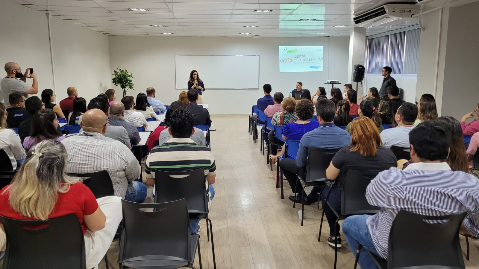 Imagem colorida em formato retangular mostra uma sala com um quadro branco e diversas cadeiras pretas ocupadas por homens e mulheres, a frente em pé, segurando um microfone está a diretora de Gestão de 1º Grau da Corregedoria, Inaiara Sá, mediadora do evento.