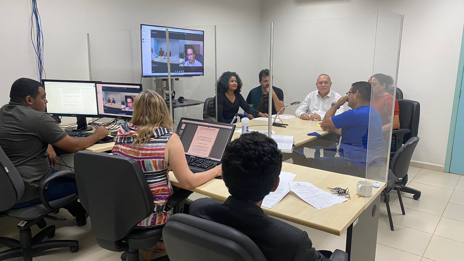 Imagem colorida em formato retangular mostra uma sala dividida com uma parede de vidro. No canto direito da foto estão dois homens e uma mulher estando dois deles mexendo em computadores que estão sobre uma mesa cor de marfim, ao fundo sentados ao redor de uma mesa estão três homens e duas mulheres, no canto superior direito está uma TV exibindo uma sala virtual.