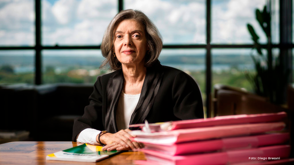 Imagem colorida da ministra Cármen Lúcia Antunes Rocha, do Supremo Tribunal Federal, sentada atrás da mesa do escritório com pastas rosas de documentos em cima da mesa e com as duas mãos em cima do livro da constituição federal. 