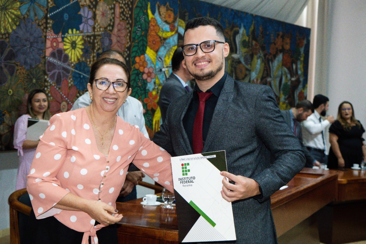 Imagem colorida em formato retangular mostra a diretora do campus Boa Vista, professora  Joseane de Souza Cortez ao lado do pós graduando Vânio José de Souza, estando este segurando o certificado que contém em sua capa a logo do IFRR.