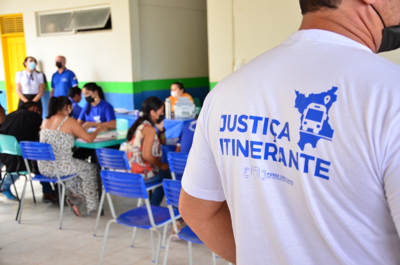  Imagem colorida em formato retangular com o fundo desfocado  mostra um servidor de costas com uma camisa branca estampada com o logotipo da vara da  justiça itinerante. 