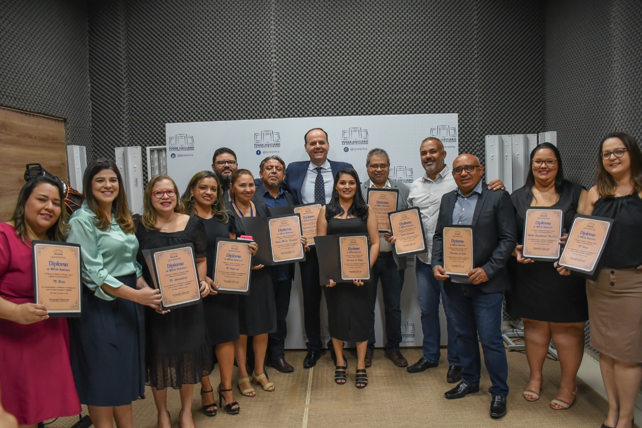 Imagem  colorida em formato retangular mostra presidente do TJRR, desembargador Cristóvão Suter ao lado dos 13 representantes dos veículos de comunicação do Estado de Roraima composto por 8 mulheres e 5 homens, os mesmos estão em pé segurando o diploma de “Mérito Judiciário” em suas mãos.