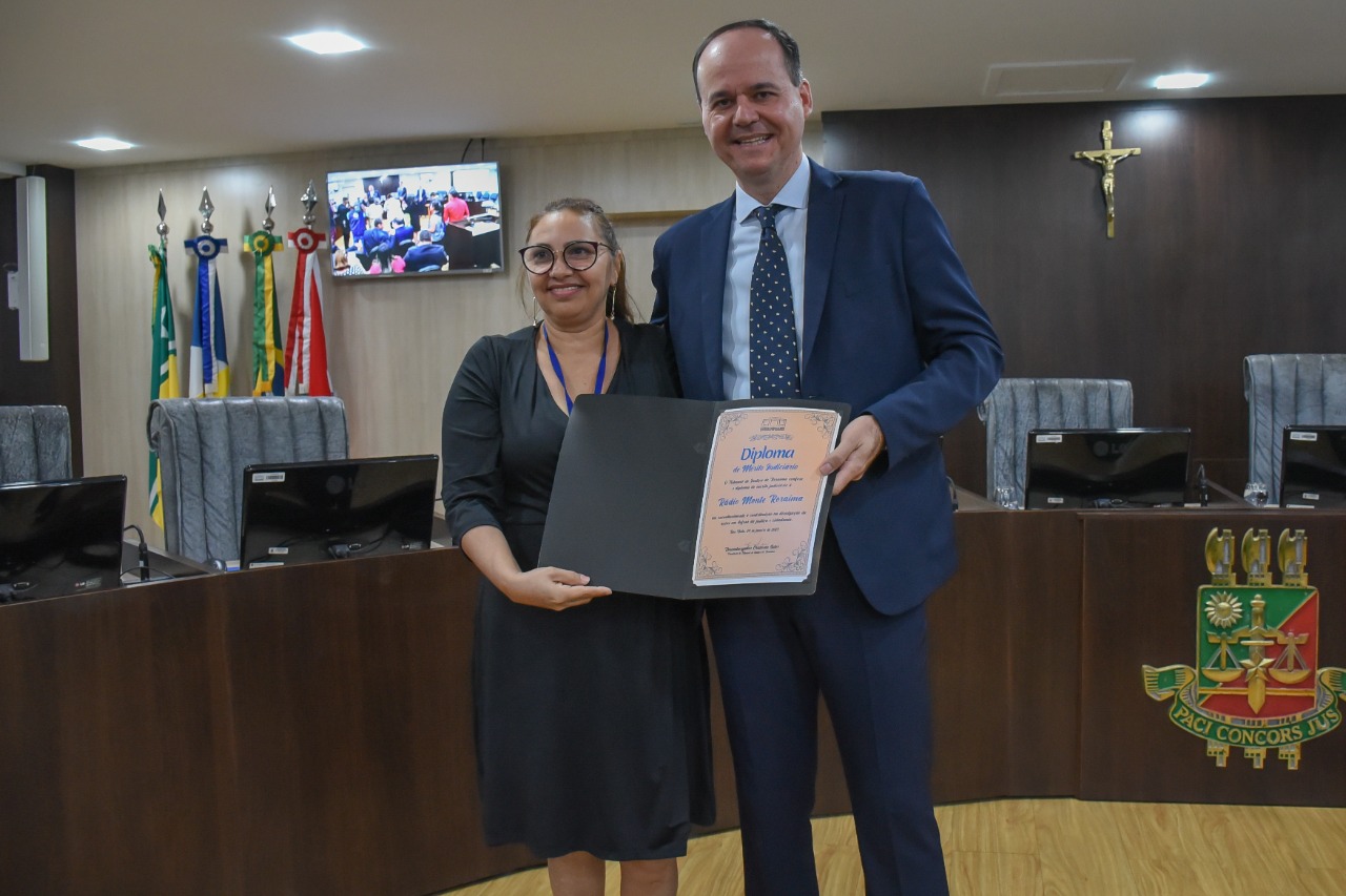 Imagem colorida em formato retangular mostra o presidente do TJRR, desembargador Cristóvão Suter ao lado da diretora administrativa da emissora Rádio Monte Roraima FM, Cléo Rocha, a  mesma segura o diploma de “Mérito Judiciário” em suas mãos.