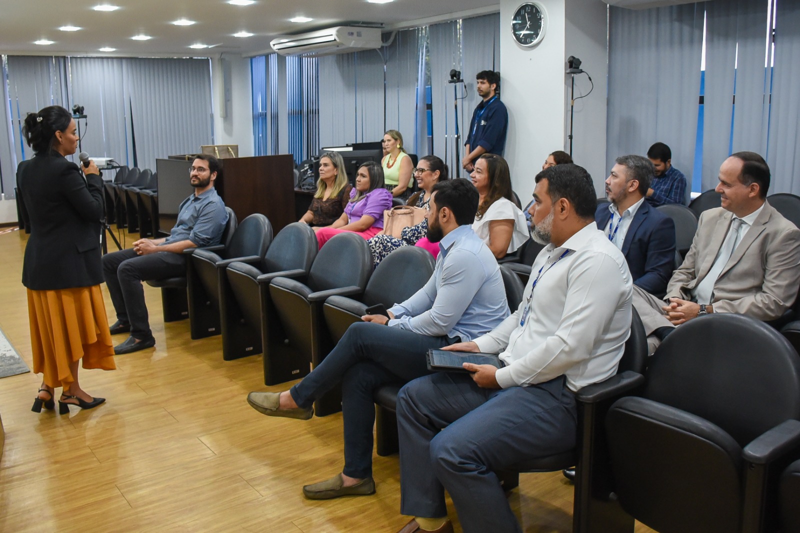 Imagem colorida em formato retangular mostra a Secretária de Gestão Estratégica (SGE), Veruska Lobo, discursando em pé com o microfone na mão para o presidente do Tribunal de Justiça de Roraima (TJRR), desembargador Cristóvão Suter, secretários e coordenadores da instituição  que estão sentados no auditório da Sala de Sessões do Palácio da Justiça.