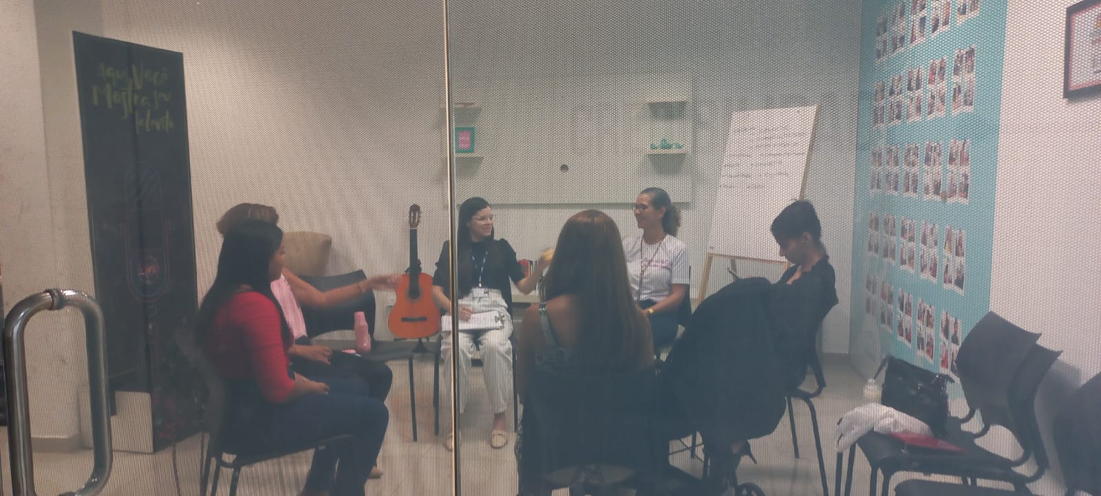 Imagem colorida em formato retangular mostra uma sala de reunião com sete mulheres sentadas em cadeiras formando um círculo, a foto foi tirada através de uma porta de vidro.