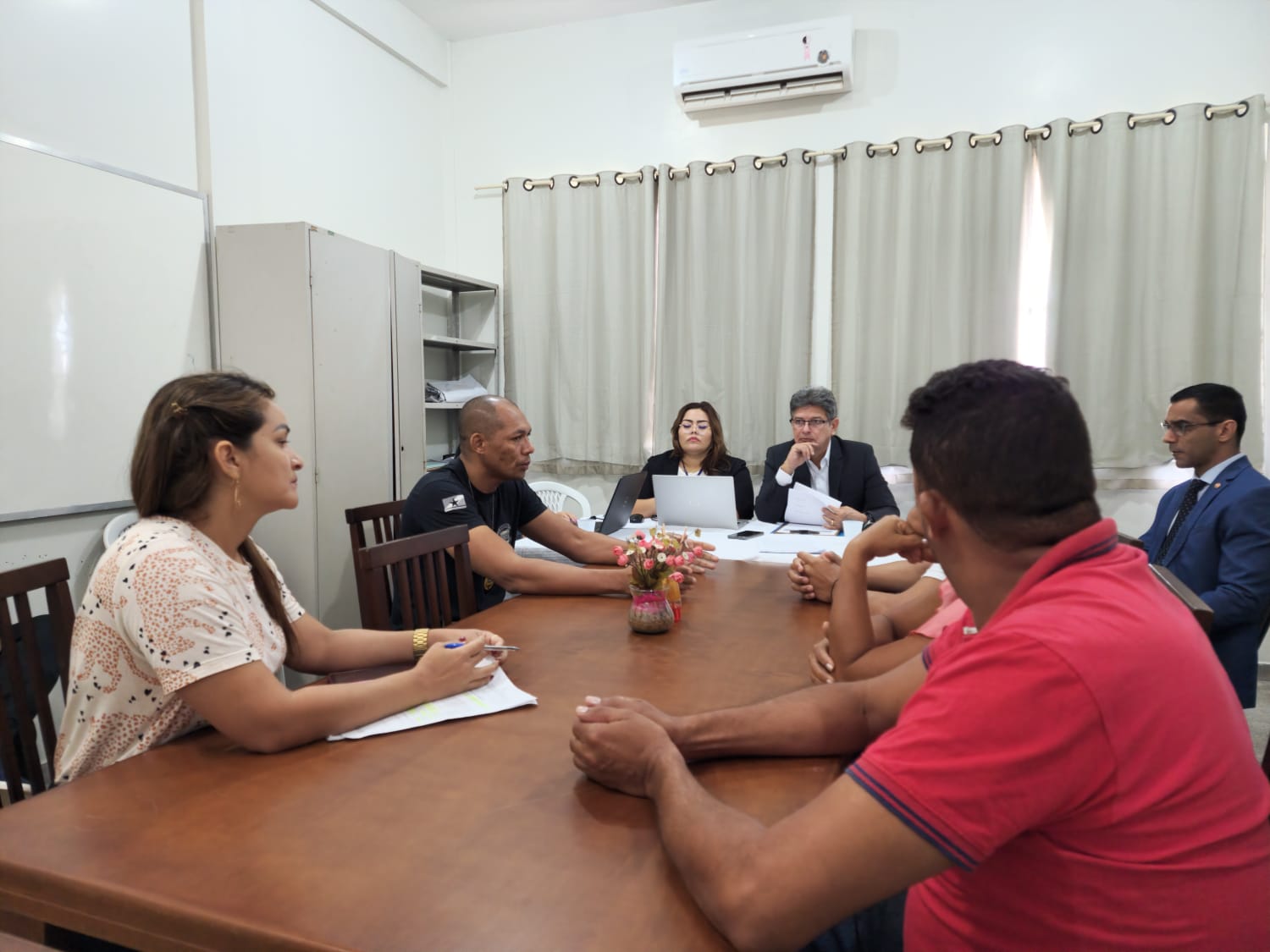 Imagem colorida mostra um grupo de oito pessoas, composto por homens e mulheres sentados ao redor de uma mesa marrom.