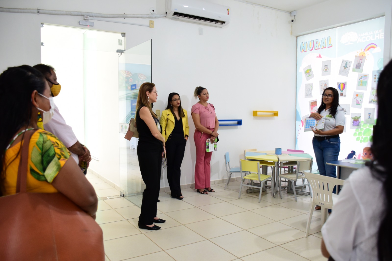 Imagem colorida mostra uma sala branca com 6 mulheres em pé observando a palestrante que está a frente mostrando um livro que está em sua mão.