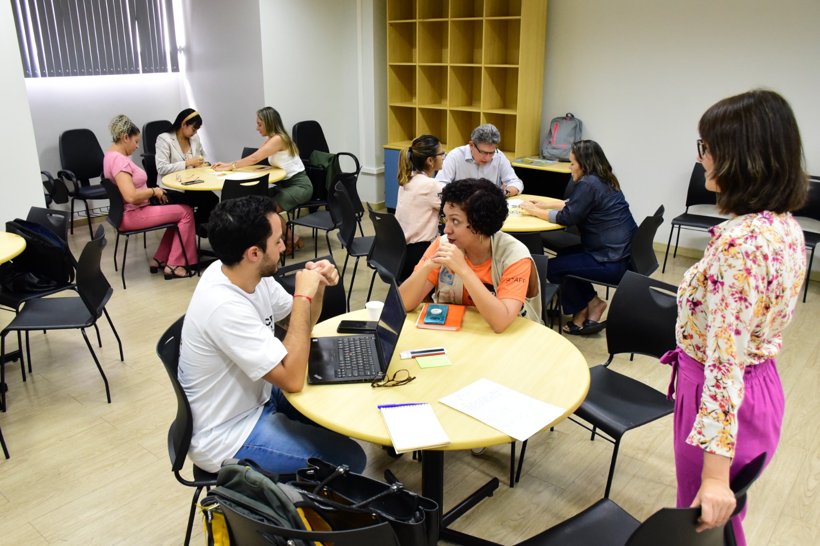 Imagem colorida mostra uma sala com 3 mesas redondas na cor marfim, com cadeiras pretas ao seu redor. duas mesas estão  ocupadas por 3 pessoas conversando, e uma está ocupada por duas pessoas, à frente dela está uma mulher em pé observando.