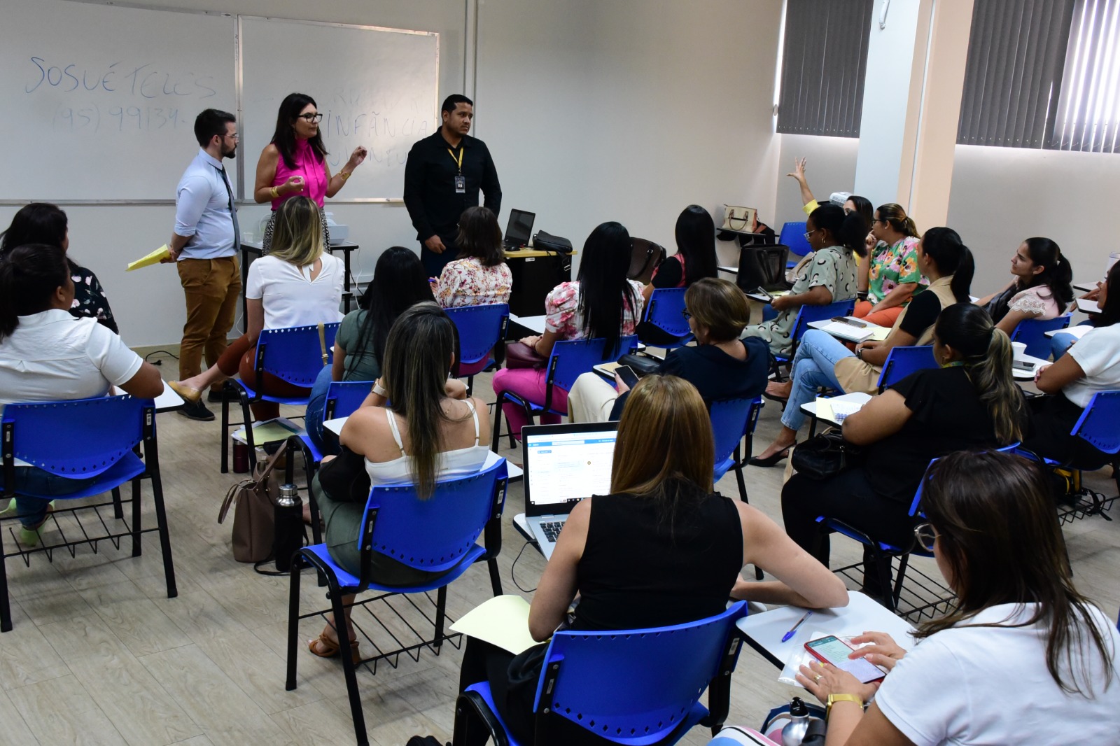 Imagem colorida mostra uma sala de aula com carteiras escolares azuis ocupadas por 18 mulheres. A frente estão três pessoas em pé, entre os quais dois homens e uma mulher.