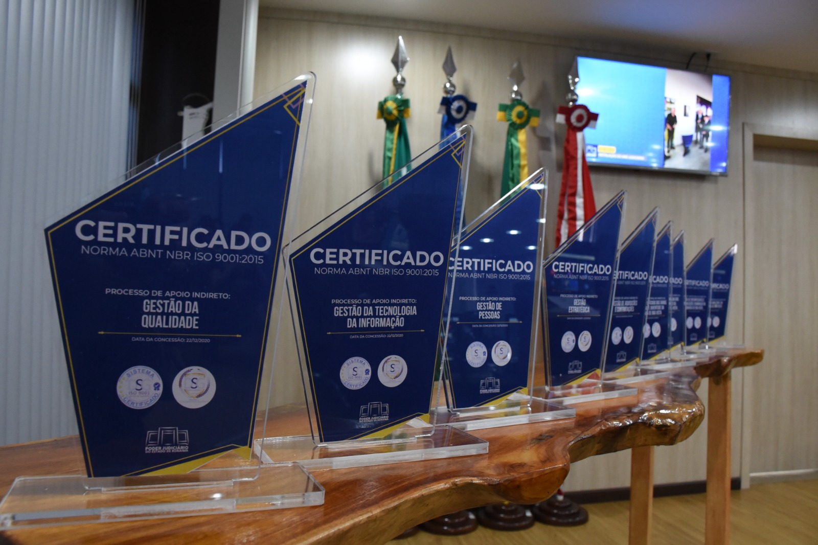 Imagem  colorida com fundo embaçado mostra uma mesa de madeira com nove troféus de certificados de qualidade de  serviços prestados ao poder público na cor azul.