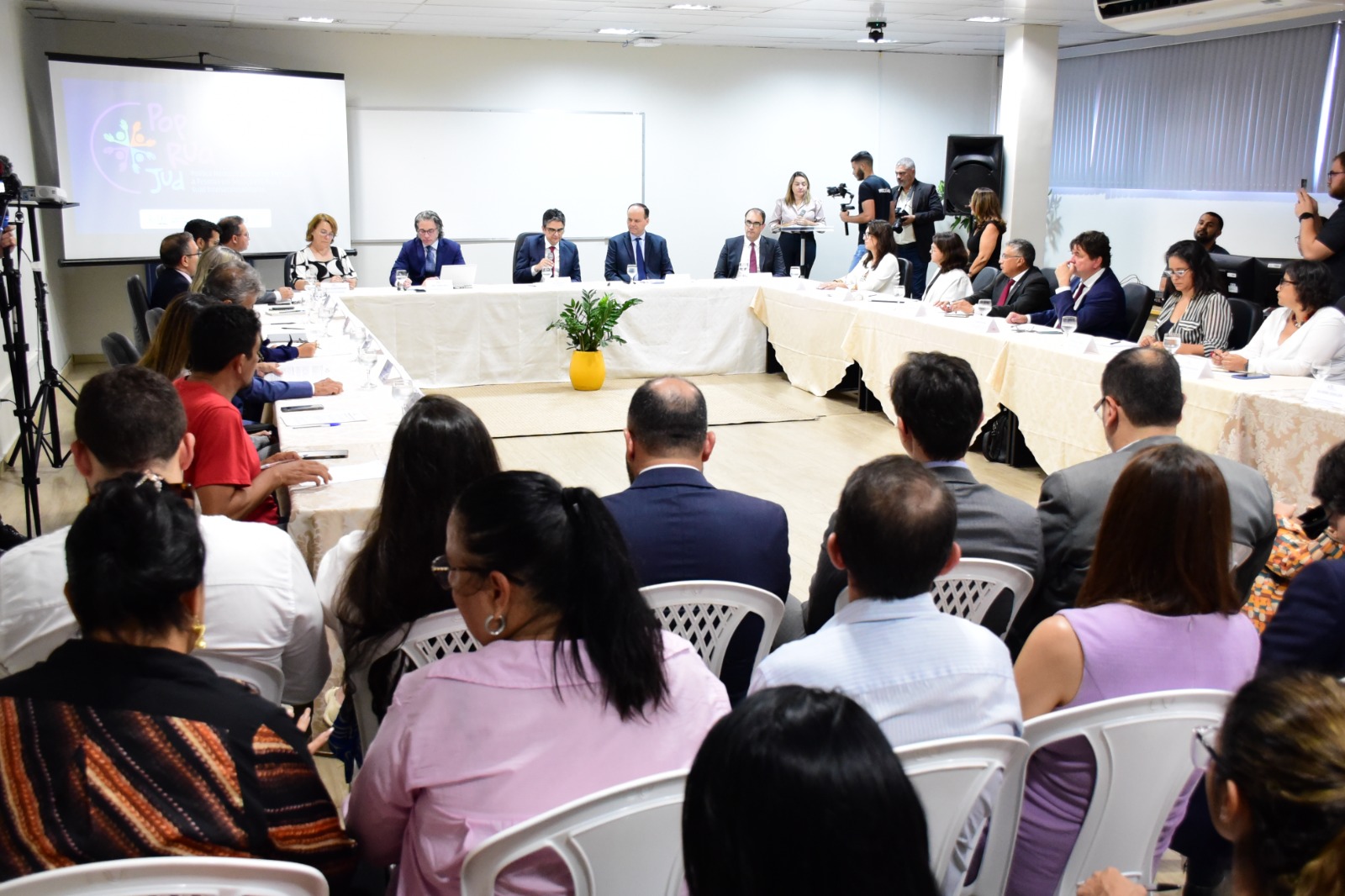 Imagem colorida mostra uma sala com cadeiras brancas ocupadas por homens e mulheres. Ao fundo, mesas cobertas com toalhas brancas posicionadas formando um retângulo, atrás estão representantes das organizações do Estado Brasileiro.