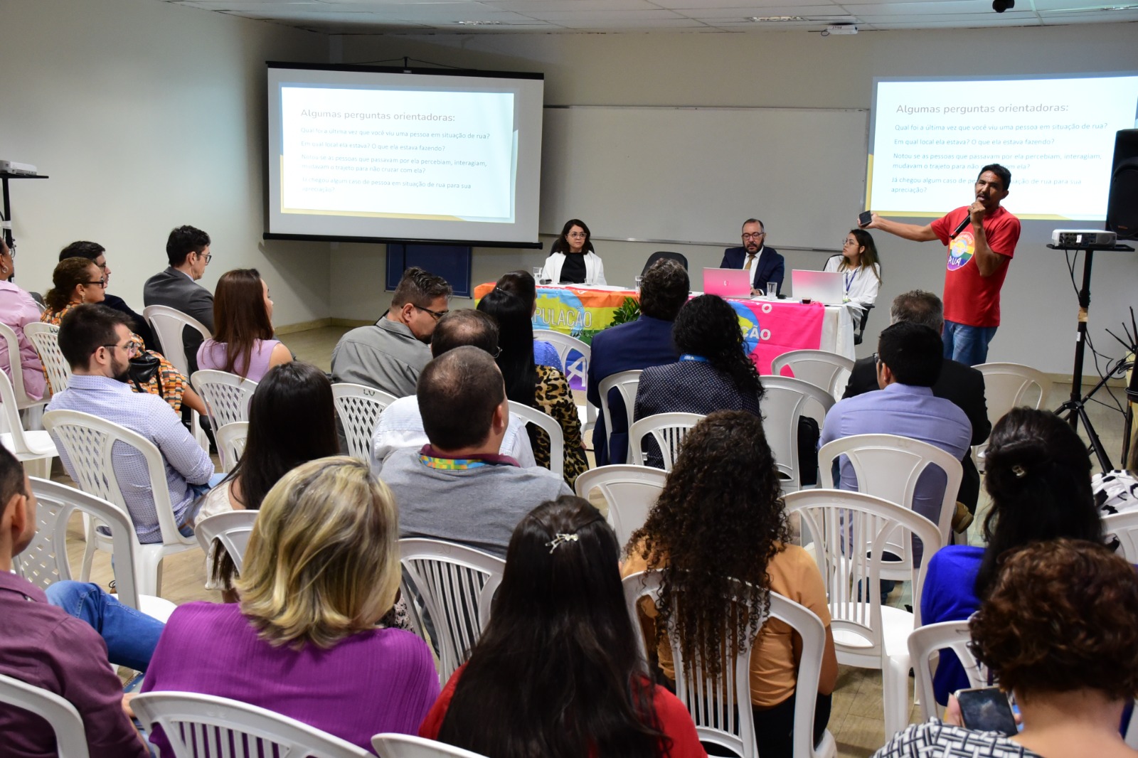 Imagem colorida mostra uma sala com cadeiras brancas ocupadas por homens e mulheres. Ao fundo, mesas cobertas com a bandeira  do Movimento Nacional População de Rua (MNPR). No canto superior esquerdo em pé, com o microfone na mão está o membro do Movimento Nacional População de Rua (MNPR), Vanilson Torres.