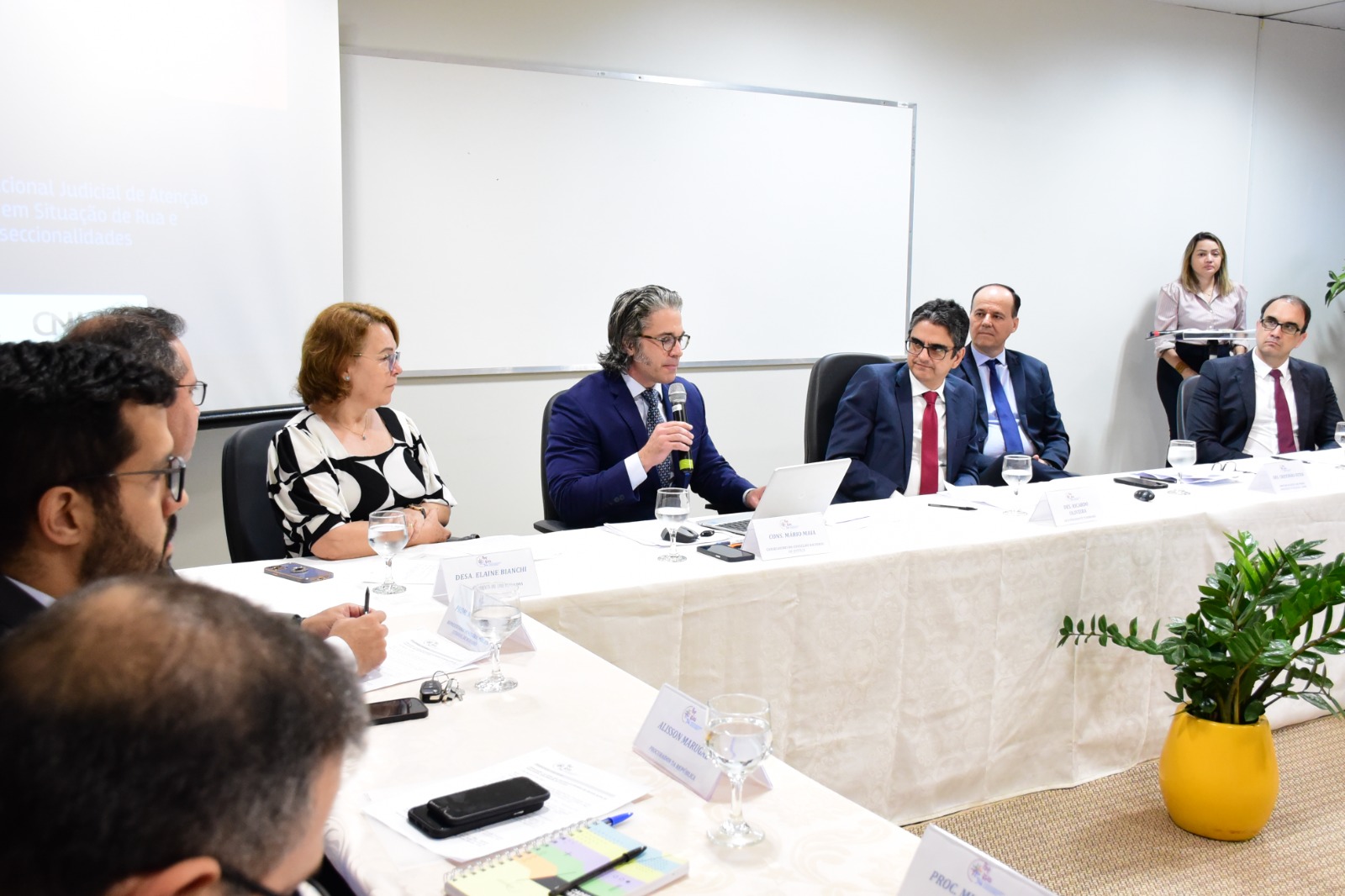 Imagem colorida mostra mesas cobertas com toalhas brancas posicionadas formando um L, atrás, em destaque estão O conselheiro do Conselho Nacional de Justiça (CNJ) presidente da Comissão Permanente de Políticas Sociais e de Desenvolvimento do Cidadão, Mário Goulart Maia com o microfone na mão, a sua direita o vice-presidente do TJRR, desembargador Ricardo Oliveira e o diretor da Escola do Poder Judiciário, desembargador Cristóvão Suter e a sua esquerda a desembargadora Elaine Bianchi