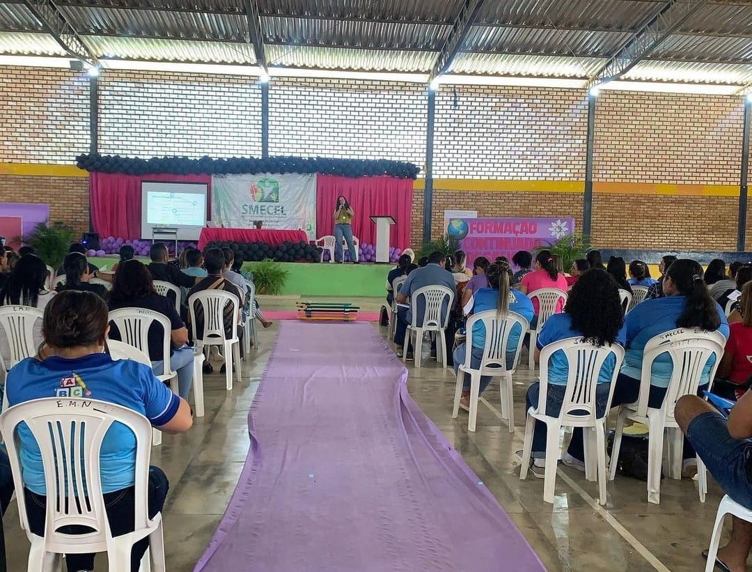 foto colorida de uma quadra chia de cadeiras d eplasticos brancas e a frente uma servidora do tjrr encima do palco palestrando