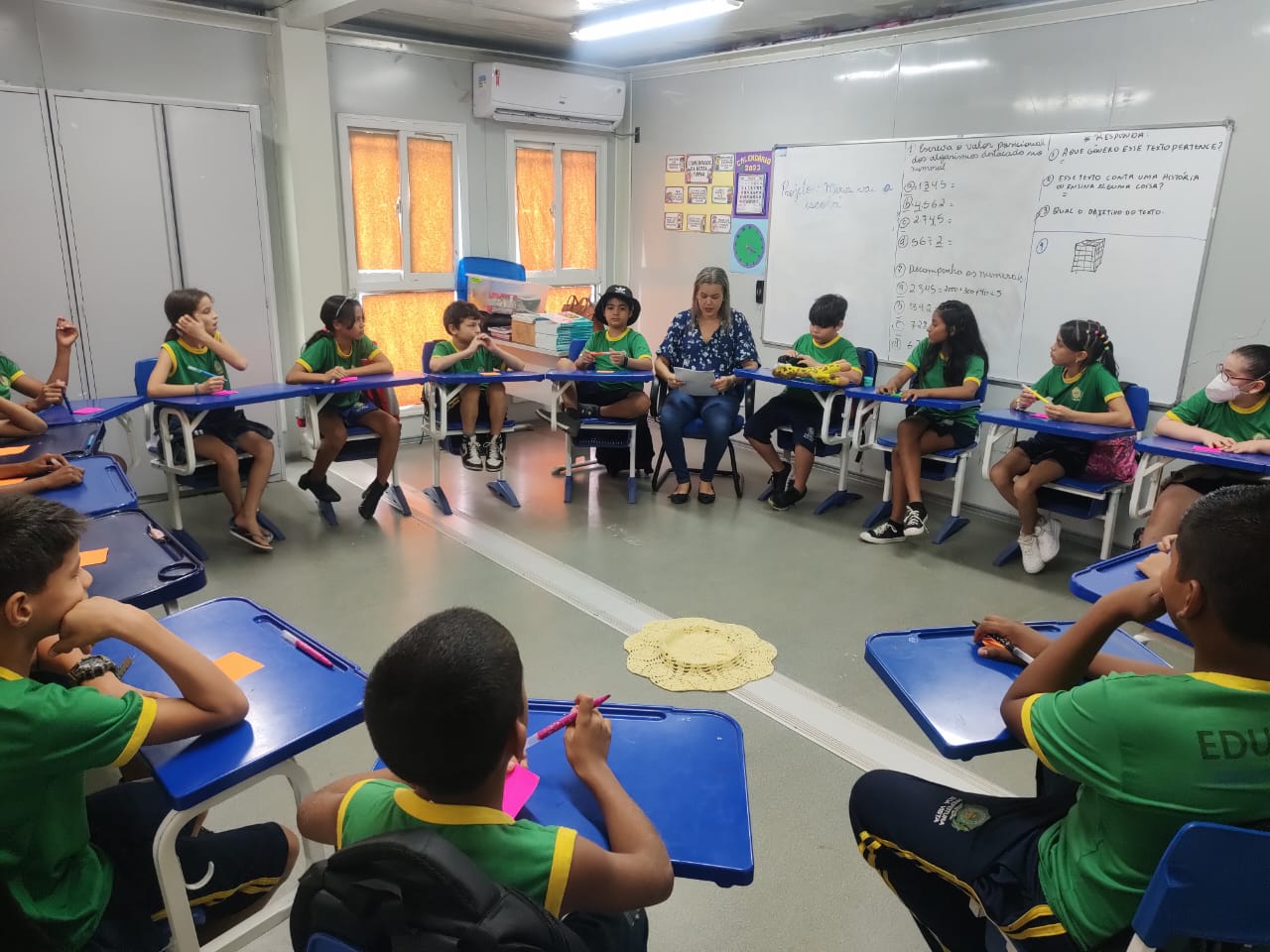 Imagem colorida mostra uma sala de aula com carteiras escolares azuis, posicionadas formando um círculo, nelas estão alunos e alunas fardados com camisas verdes com golas amarelas.