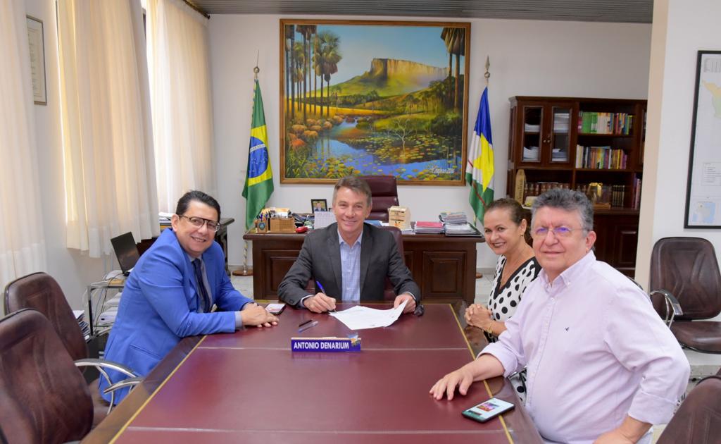 Imagem colorida mostra uma sala com uma mesa de reunião ocupada pelo o presidente do Tribunal de Justiça de Roraima, desembargador Jésus Nascimento, o  governador Antonio Denarium, o chefe da Casa Civil, Flamarion Portela e a juíza Graciete Sotto Mayor. No fundo, um quadro de uma paisagem representando o Monte Roraima. Ao lado do quadro estão hasteadas as  bandeiras do Brasil e do Estado de Roraima.