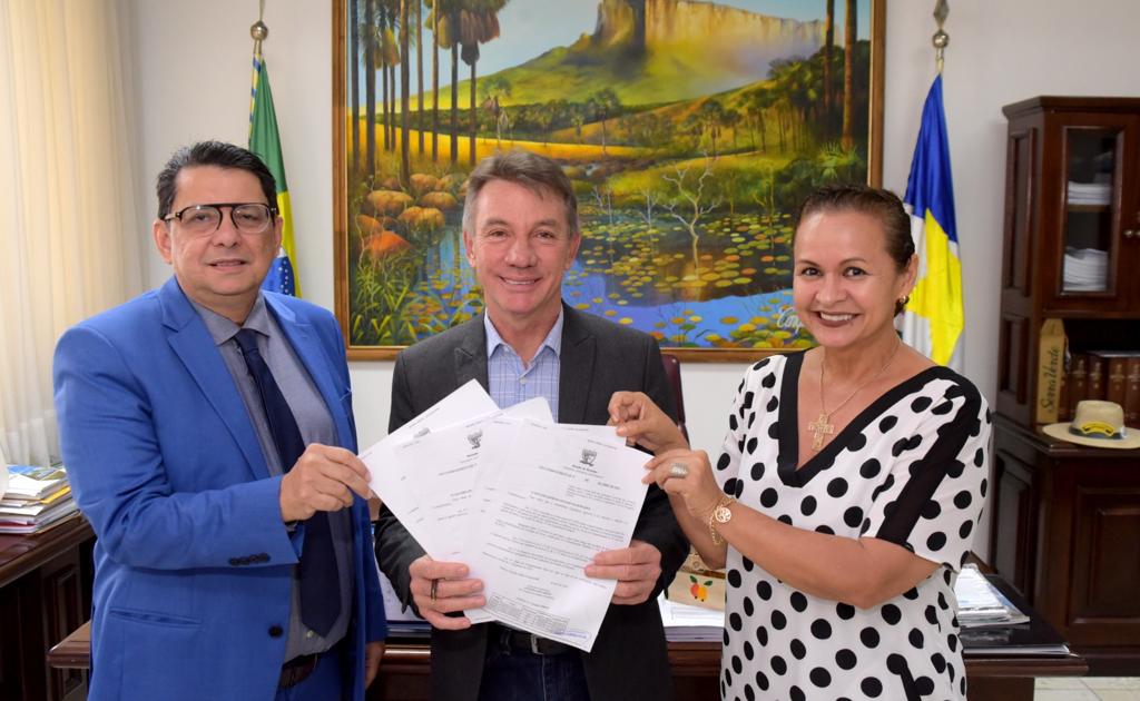 Imagem colorida mostra o presidente do Tribunal de Justiça de Roraima, desembargador Jésus Nascimento, tocando nos papéis segurados pelo  governador Antonio Denarium, a mesma ação é reproduzida pela  juíza Graciete Sotto Mayor. No fundo, um quadro de uma paisagem representando o Monte Roraima. Ao lado do quadro estão hasteadas as  bandeiras do Brasil e do Estado de Roraima.