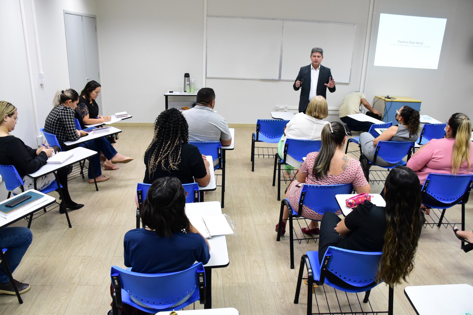 magem colorida mostra uma sala de aula com homens e mulheres sentados em carteiras escolares nas cortes azul e branca. A frente Conduzindo a reunião está o juiz titular da 1ª Vara da Infância e Juventude, Parima Dias Veras