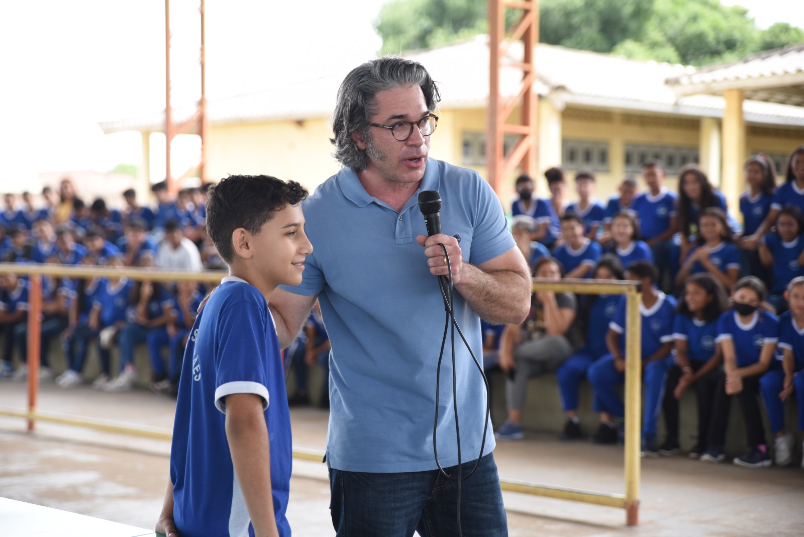 Imagem colorida com o fundo embaçado, mostra uma quadra de esporte com alunos e alunas da Escola Estadual Ulysses Guimarães. No centro está o presidente da Comissão Permanente de Políticas Sociais e de Desenvolvimento do Cidadão, Mário Goulart Maia, conselheiro do CNJ, segurando um microfone e colocando a mão sobre o ombro de um adolescente.