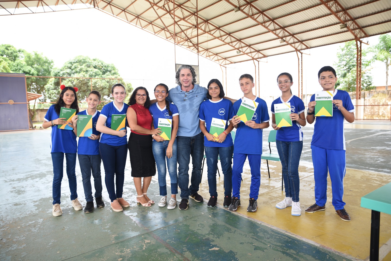 Imagem colorida mostra uma quadra de esporte. No centro estão o presidente da Comissão Permanente de Políticas Sociais e de Desenvolvimento do Cidadão, Mário Goulart Maia, conselheiro do CNJ, e a gestora Isane Pedrosa, posando para a foto ao lado de cinco alunas e três alunos que seguram o livro da constituição.