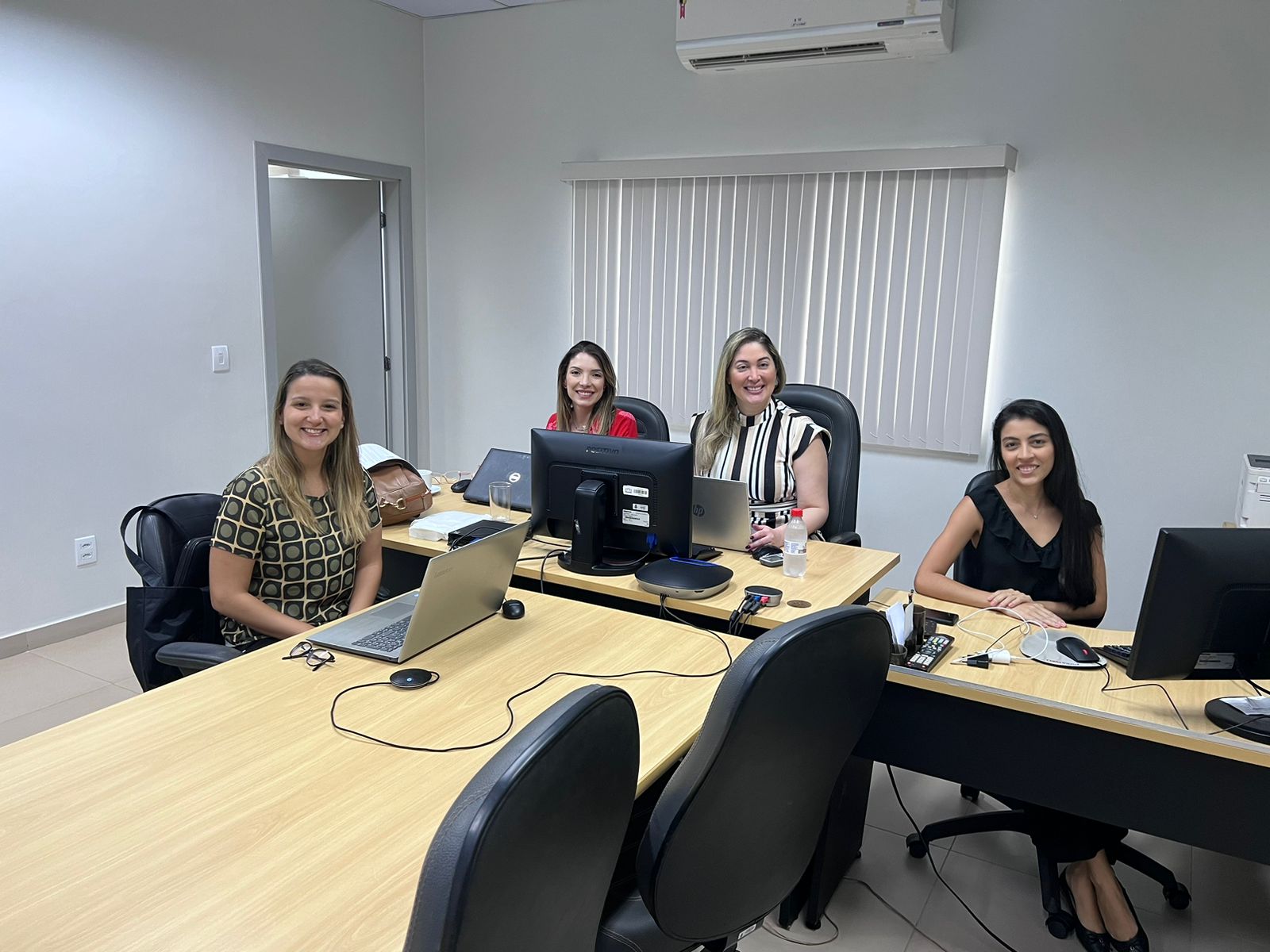 Imagem colorida mostra uma sala de reunião com mesas na cor marfim com computadores e notebook sobre elas. Ao redor da mesa, sentadas,  estão a juíza presidente da sessão, Rafaella Holanda Silveira, a promotora de Justiça Lara Von Held Cabral Fagundes, a defensora pública Elisa Rocha Teixeira Netto e a oficial de gabinete, Letícia Machado, sorrindo, posando para a foto.