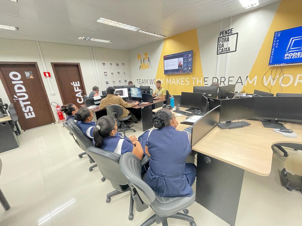 Imagem colorida mostra a sala do Núcleo de Comunicação e Relações Institucionais (NUCRI), com servidores, estagiários e quatro  colaboradoras assistindo uma reunião virtual exibida em uma smart TV.