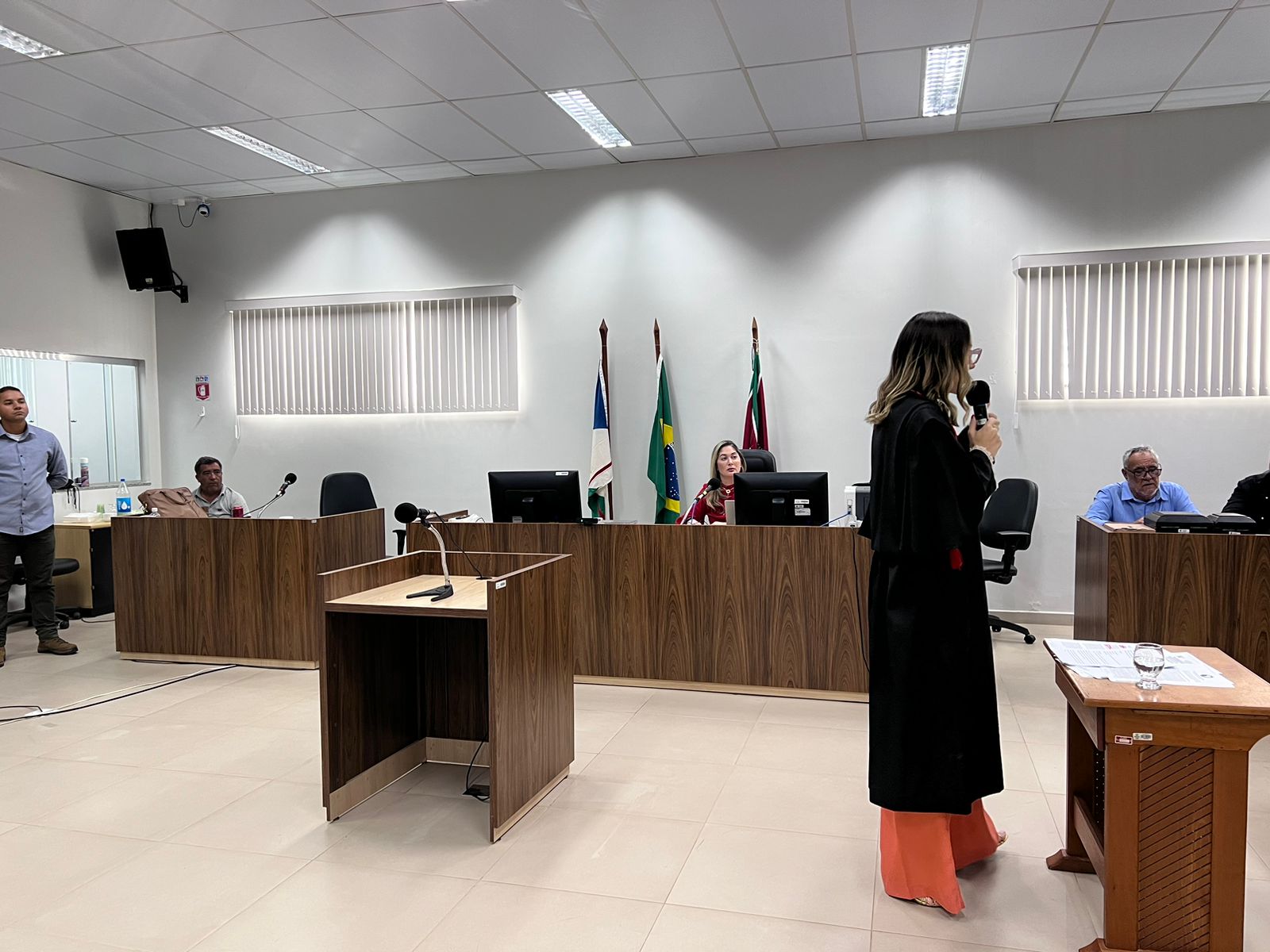 Imagem colorida mostra uma sessão de julgamento do Tribunal do Júri na  Comarca do Município de São Luiz. No lado esquerdo da foto está uma magistrada usando toga , posicionada lateralmente com o microfone na mão.