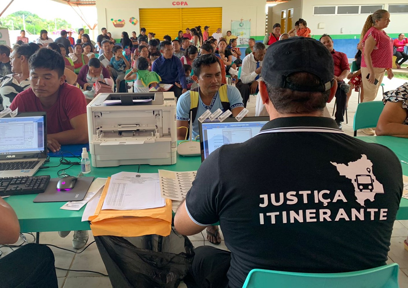 Imagem colorida mostra um servidor de costa vestindo uma camisa preta com a logo da Justiça itinerante . Ao fundo, homens e mulheres aguardando sentados a espera pelo atendimento.