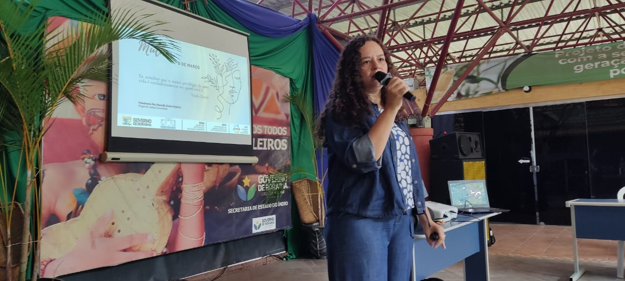 Imagem colorida mostra a coordenadora do Programa Justiça Comunitária, Marcelle Grécia Wottrich, em pé segurando um microfone na mão durante a palestra.