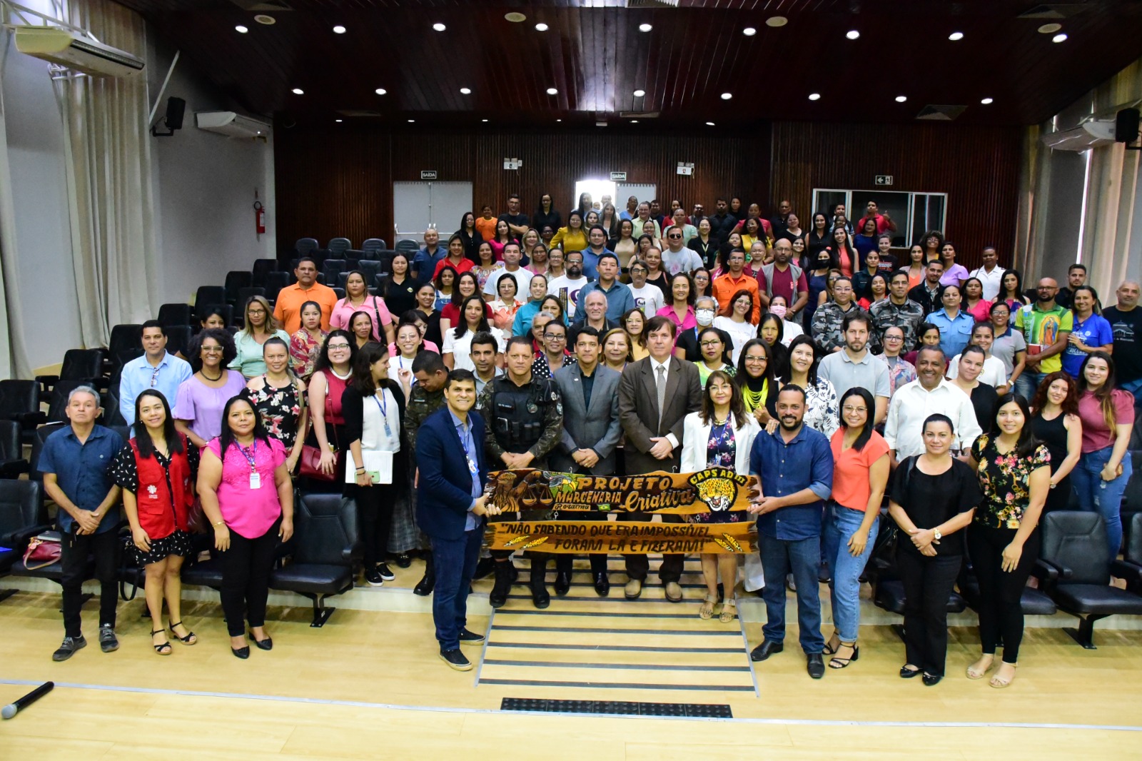 Imagem colorida mostra o juiz titular da Vepema, Alexandre Magno posando e os representantes de escolas municipais e estaduais, e no segundo dia, entidades públicas (saúde, segurança, assistência social, ambiental), privadas (ONGs, associações), filantrópicas (igrejas, grupos de mútua ajuda, comunidades terapêuticas), em pé posando para a foto. a frente estão dois homens segurando uma placa de madeira entalhada com palavras e desenhos.