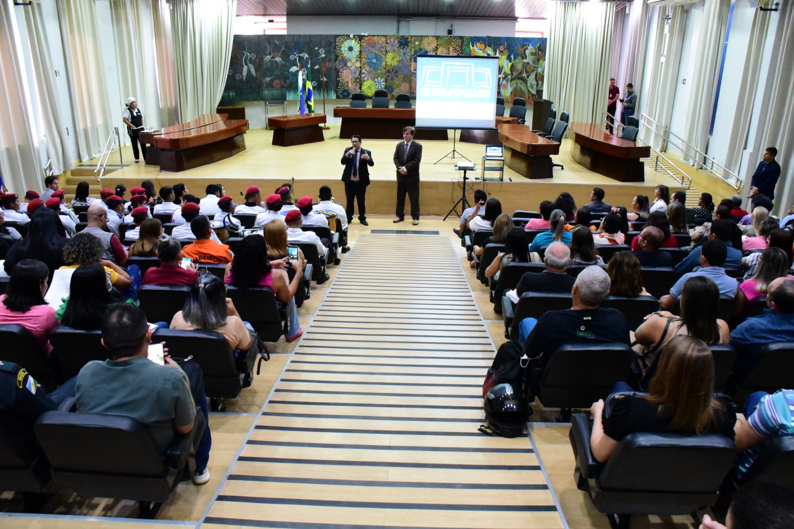 Imagem colorida mostra o auditório do Fórum Cível lotada com representantes de escolas municipais e estaduais, e no segundo dia, entidades públicas (saúde, segurança, assistência social, ambiental), privadas (ONGs, associações), filantrópicas (igrejas, grupos de mútua ajuda, comunidades terapêuticas). A frente o presidente do Tribunal de Justiça de Roraima, desembargador Jésus Nascimento, discursando com o microfone na mão ao lado do juiz titular da Vepema, Alexandre Magno.