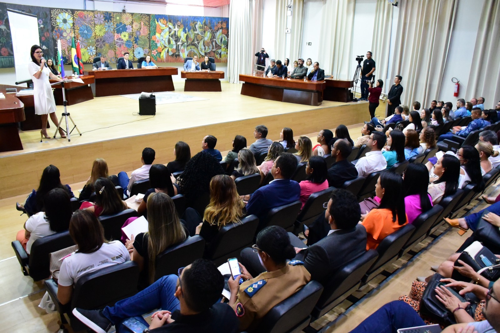 Imagem colorida mostra o auditório do Fórum Sobral Pinto lotado de servidoras, servidores e colaboradores do poder Judiciário. A frente quatro mesas de madeiras ocupadas por autoridades. No canto superior esquerdo, em pé segurando um microfone está a juíza auxiliar da Presidência do CNJ, Fabiane Pieruccini.