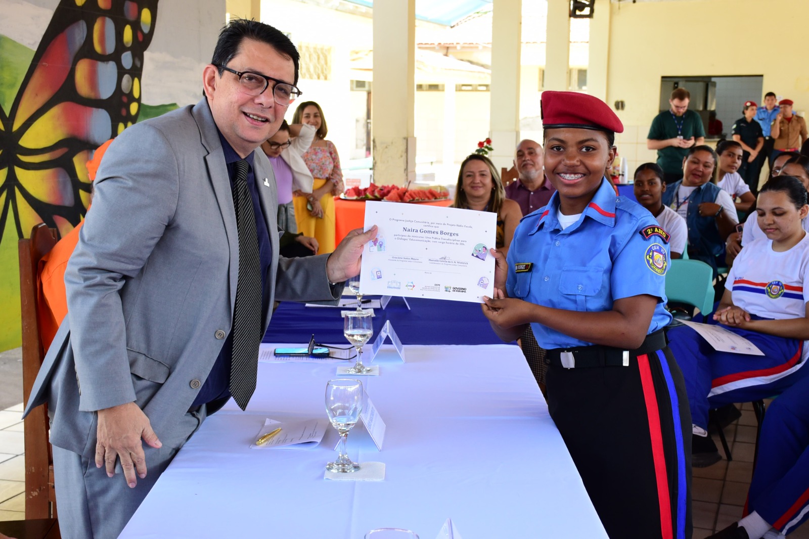 Imagem colorida mostra o presidente do Tribunal de Justiça de Roraima, desembargador Jésus Nascimento tocando o certificado ao mesmo tempo que uma aluna uniformizada  da Escola Estadual Militarizada Maria dos Prazeres Mota. 