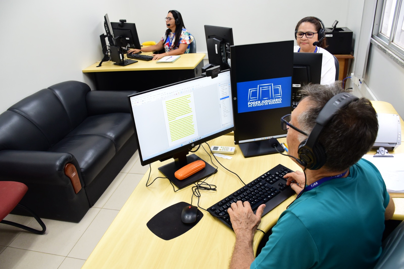 Imagem colorida mostra uma sala com um homem e duas mulheres trabalhando em mesas. Sobre as mesas estão acessórios e equipamentos de informática como webcams, headsets e scanners. No canto esquerdo da imagem há um sofá preto de dois lugares.