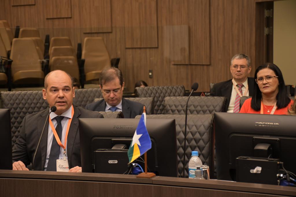  Imagem colorida mostra o juiz Eduardo Álvares Carvalho, coordenador do Centro Judiciário de Solução de Conflitos e Cidadania do Tribunal de Justiça de Roraima (CEJUSC/TJRR),  durante debate. A frente está o microfone e uma pequena bandeira do estado, ao fundo estão dois homens e uma mulher sentados em cadeiras pretas.