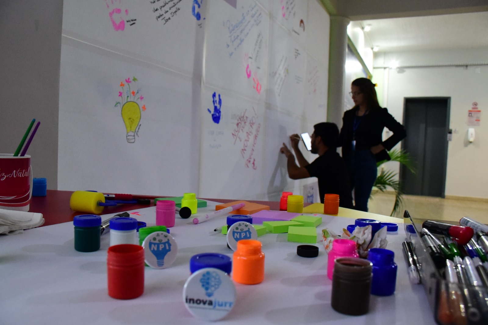  Imagem colorida mostra uma mesa com diversos potes de tinta guache, post-its e canetas coloridas. Ao fundo há um homem agachado escrevendo em  uma parede com cartolinas brancas decoradas com mãos e frases, ao seu lado, em pé, está uma mulher observando a ação.