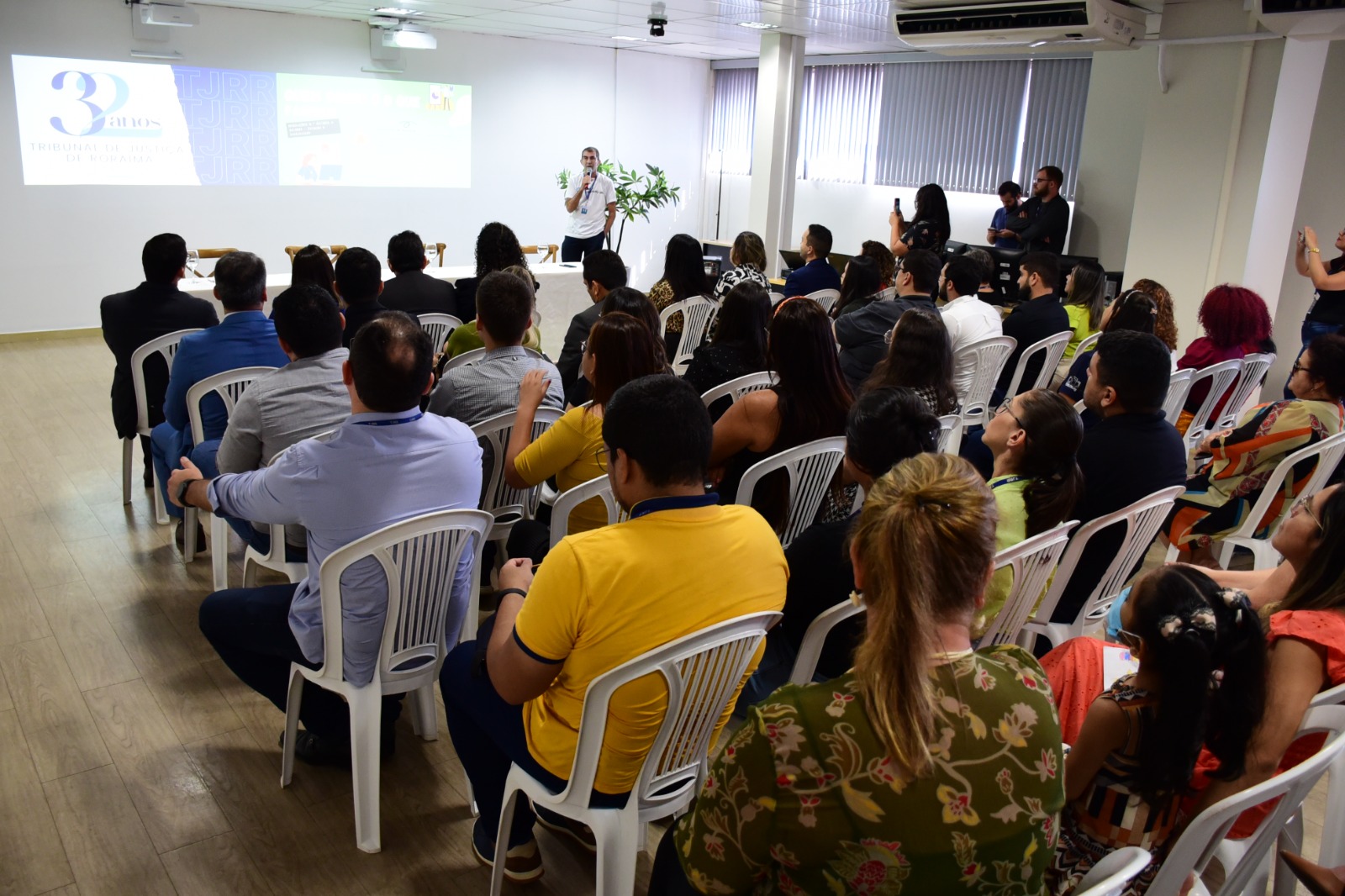 FOTO COLORIDA DE UM AUDITORIO CHEIO DE GENTE SENTADO EM CADEIRAS DE PLASTICO